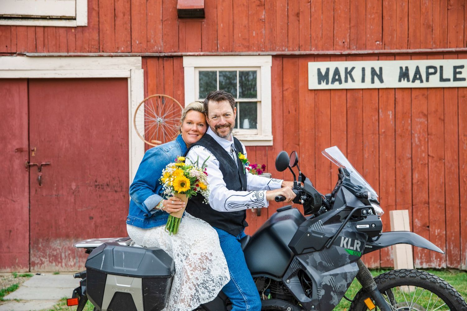 Smiling couple on a black motorcycle in front of a red barn, bride holding sunflowers and bouquet