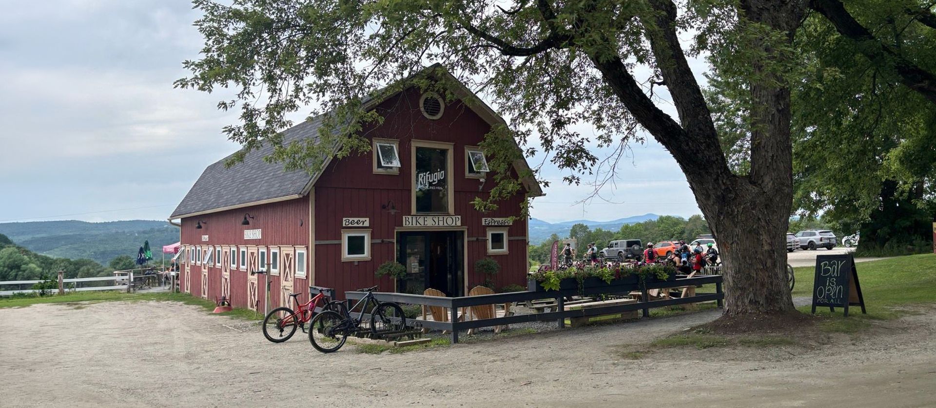 Red barn-style building with bicycles, a tree, and people seated outdoors by a lake