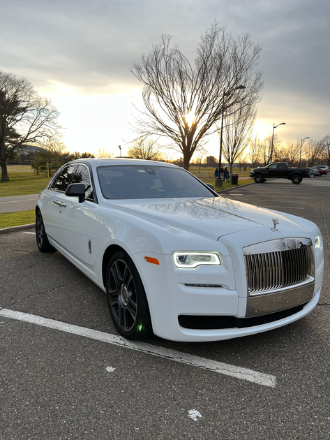 A white Rolls-Royce parked on an asphalt lot during a sunset with trees in the background.