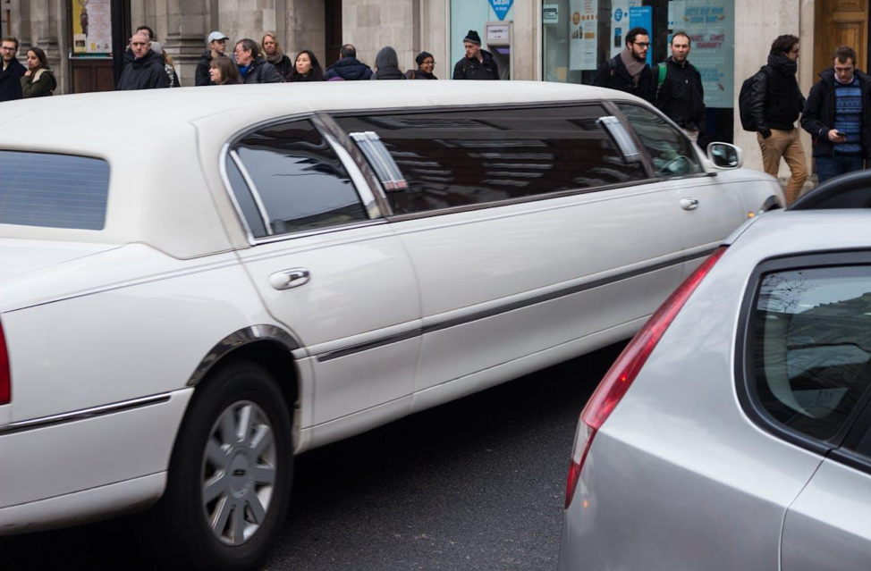 A white stretch limousine parked on a city street with people standing on the sidewalk in the background.