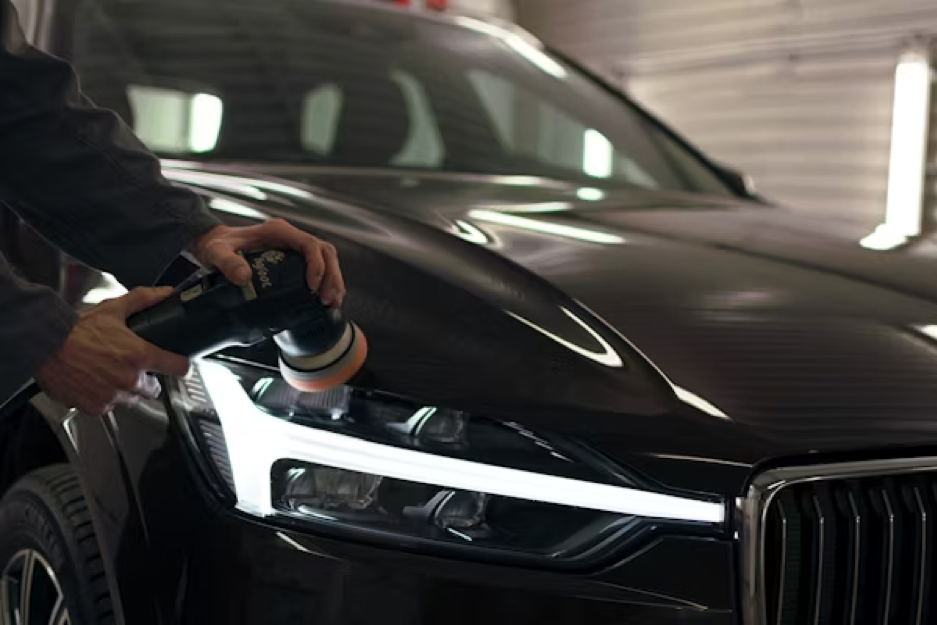A person uses an electric buffer to polish the hood of a black SUV in a workshop.