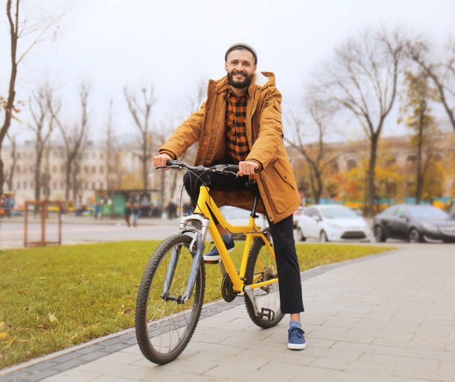 Smiling man in a mustard coat rides a yellow bicycle on a paved park path in autumn