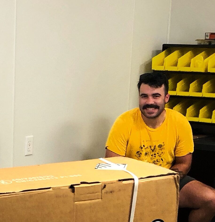 Smiling man in a yellow shirt sitting behind a large cardboard box in a room with yellow storage bins
