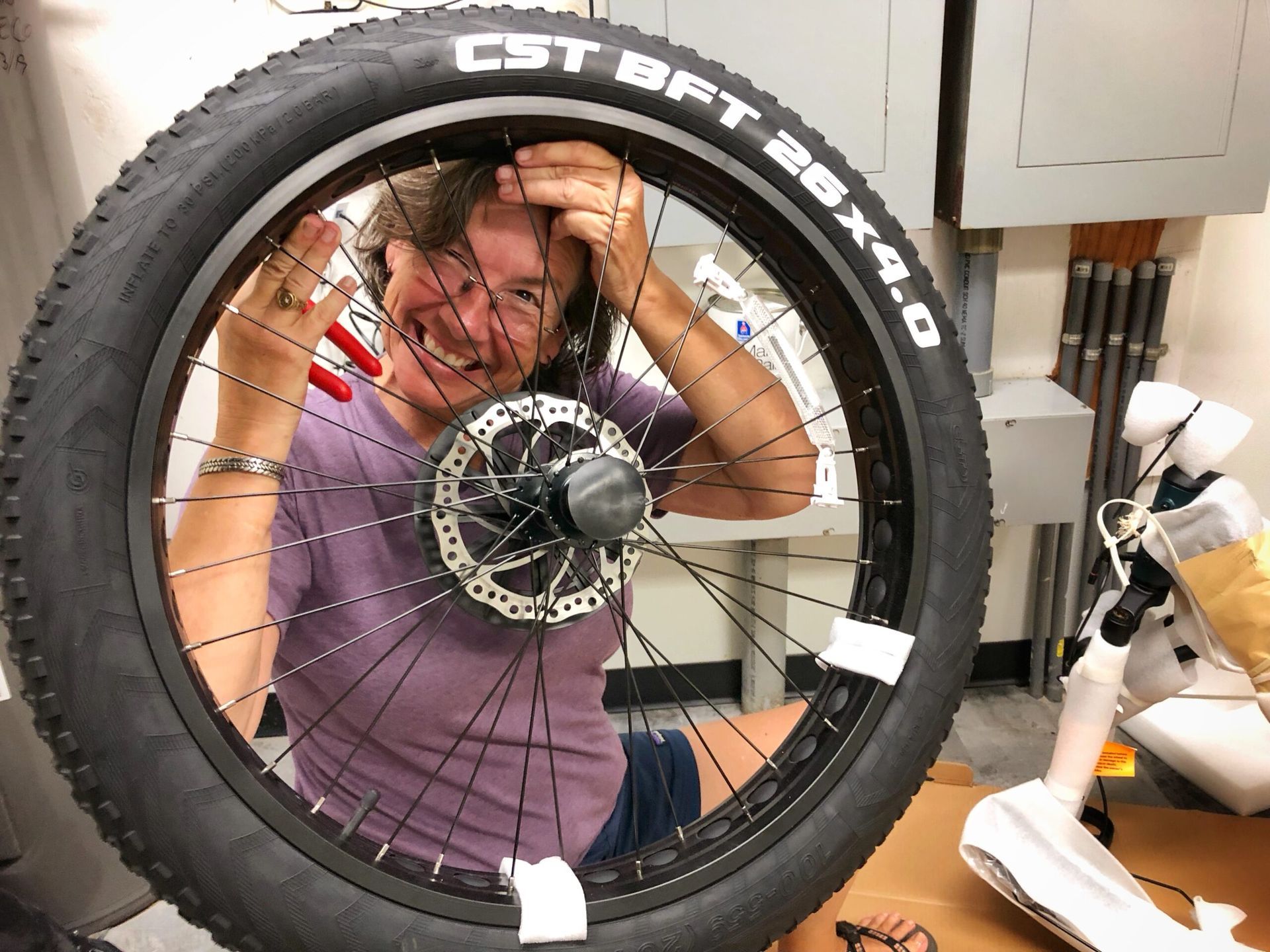 Person smiling behind a bicycle wheel, with a white robot in a workshop.