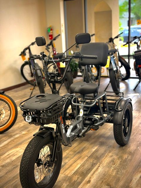 Three-wheeled black electric mobility scooter with seat indoors, with bicycles in the background