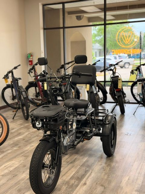 Black three-wheeled electric trike displayed indoors among bicycles in a showroom.