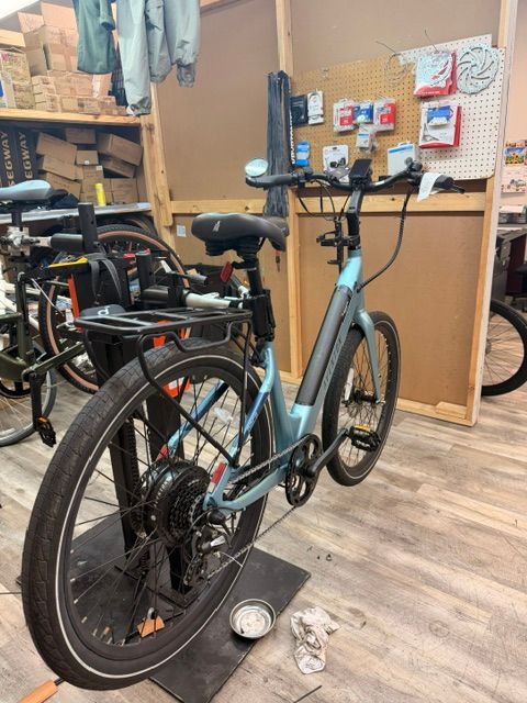 Gray e-bike on a stand in a bike workshop, with tools, shelves, and a corkboard in the background