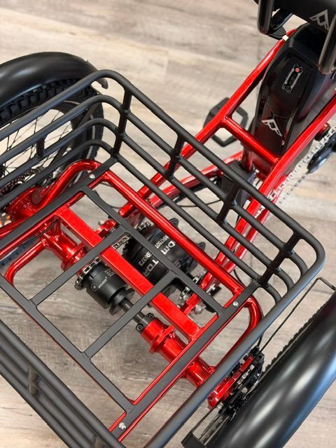 Red-framed cargo tricycle with black front rack and wheels, viewed from above on a wooden floor