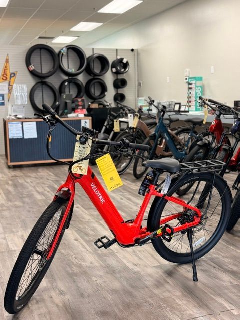 Red e-bike displayed in a bicycle shop, with tires and other bikes in the background