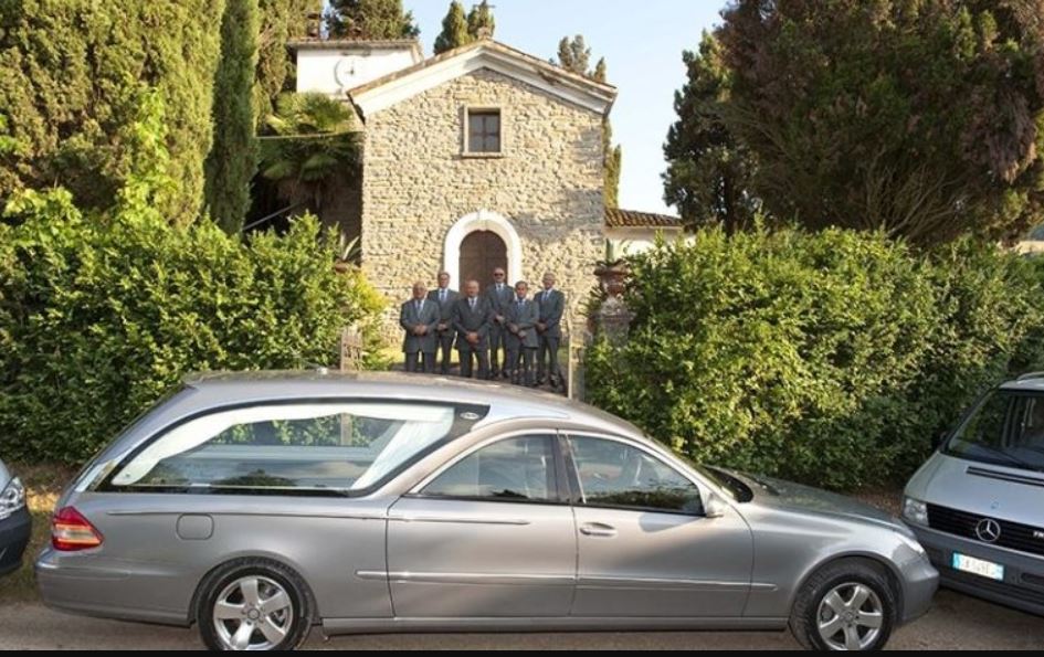 Un carro funebre parcheggiato davanti a un edificio in pietra, con diversi uomini in abito scuro in piedi nelle vicinanze.