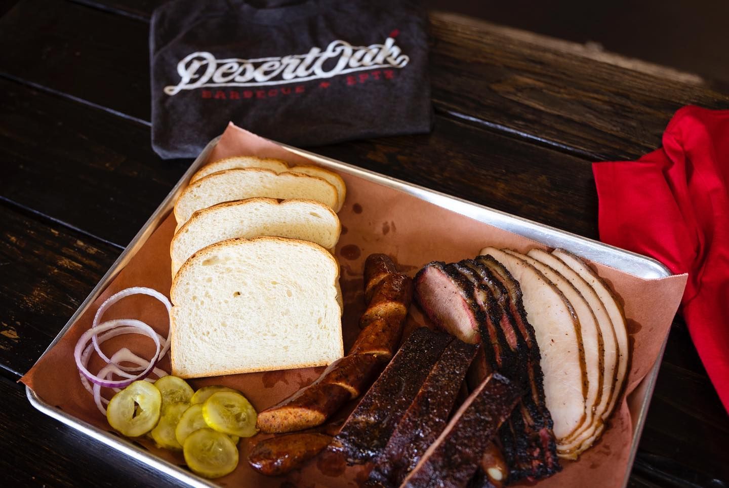 Platter of sliced smoked meats, bread, pickles, and onions on a rustic table with a red napkin
