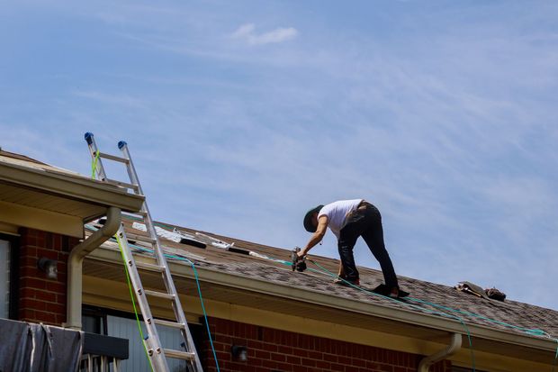 Worker repairing a shingled roof beside a ladder under a blue sky
