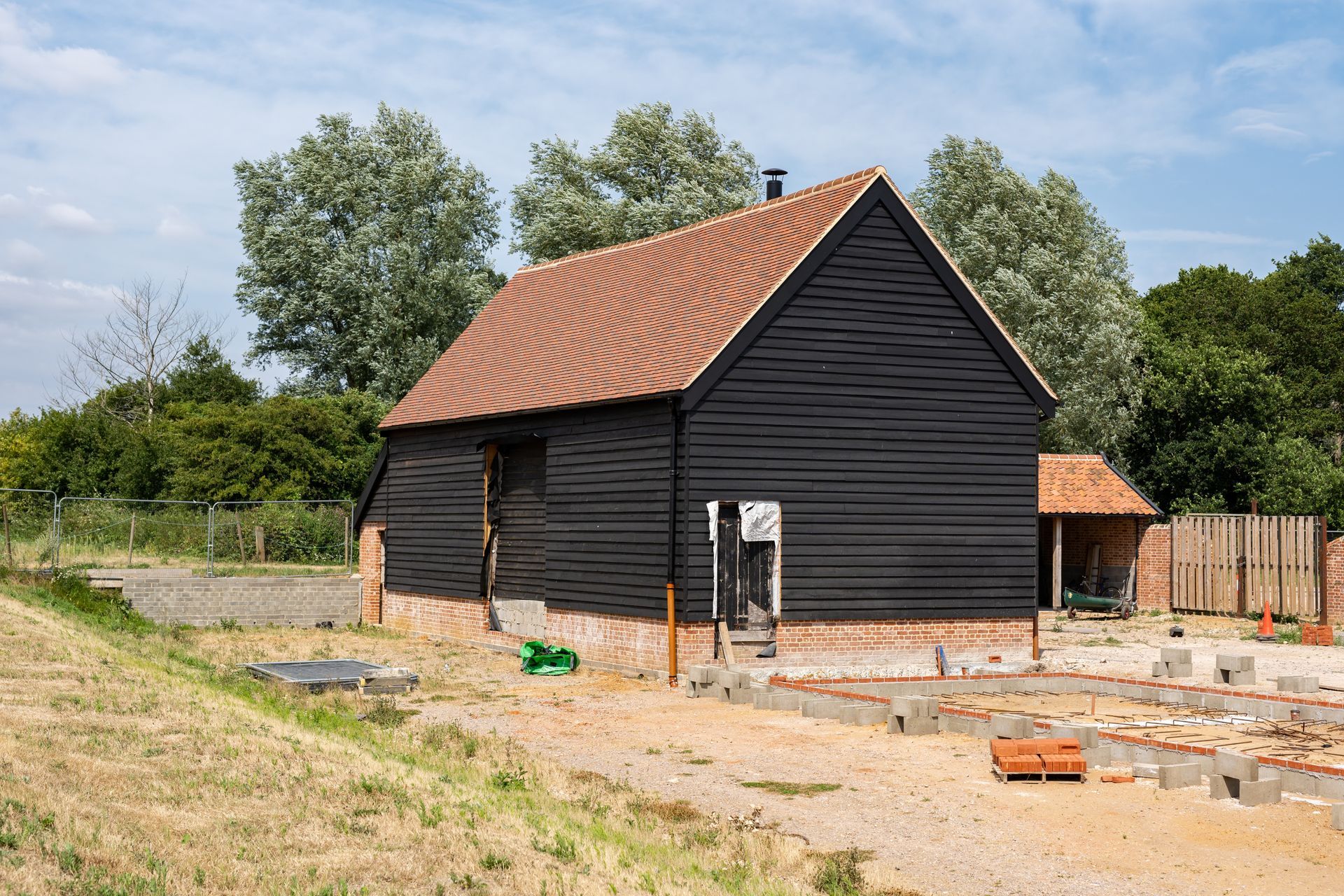 Black wooden house with red roof under construction on a dirt lot, surrounded by trees.