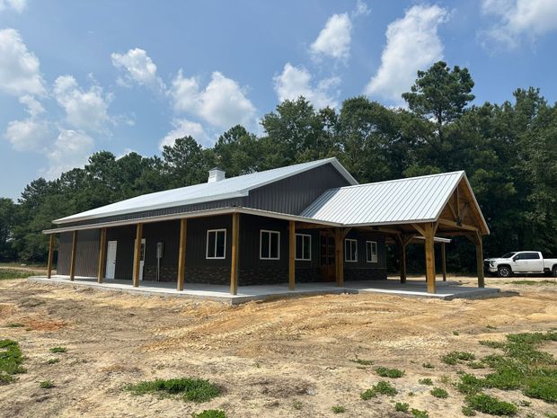 New metal-roofed wooden building under construction on a dirt lot with trees and a parked car nearby