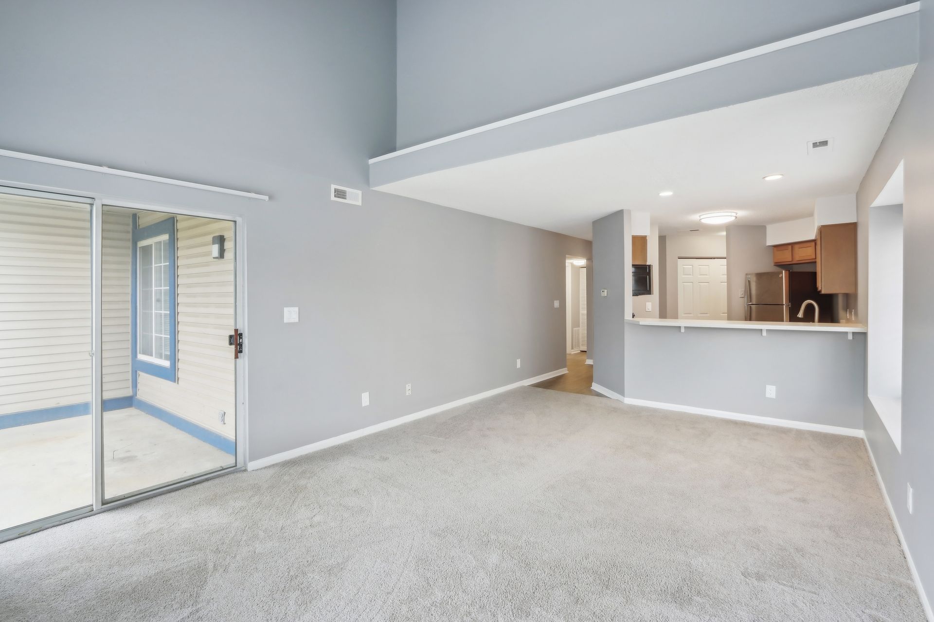 Empty living room with gray walls, carpet, and sliding glass door. Kitchen visible.