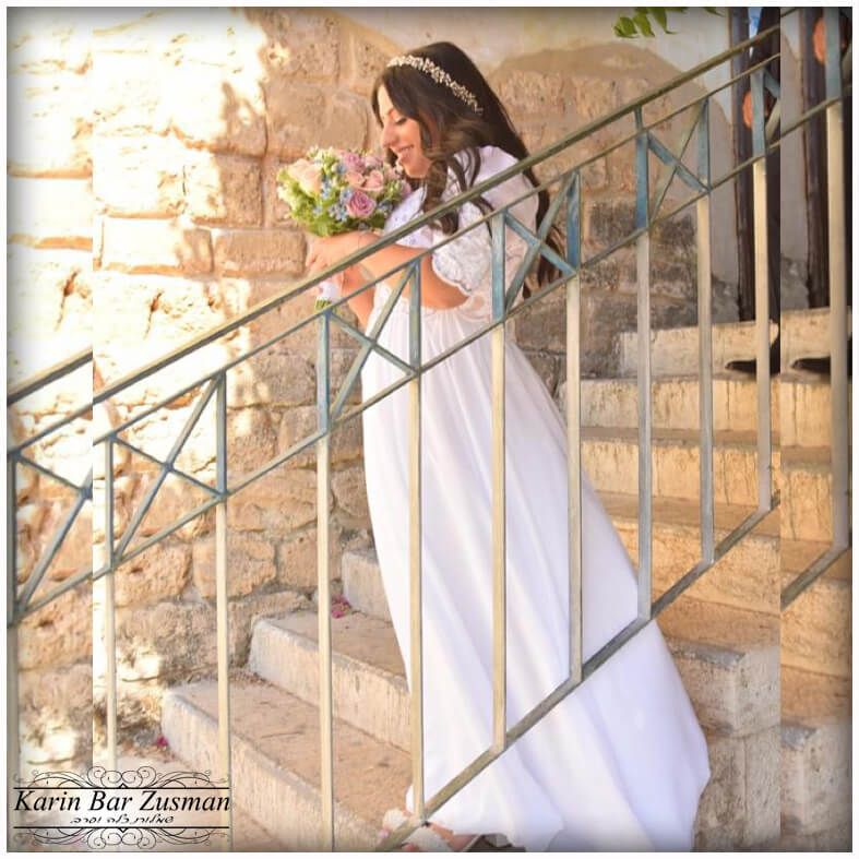 כלה בשמלה לבנה, אוחזת זר פרחים, מחייכת בזמן שהיא עומדת על מדרגות אבן.