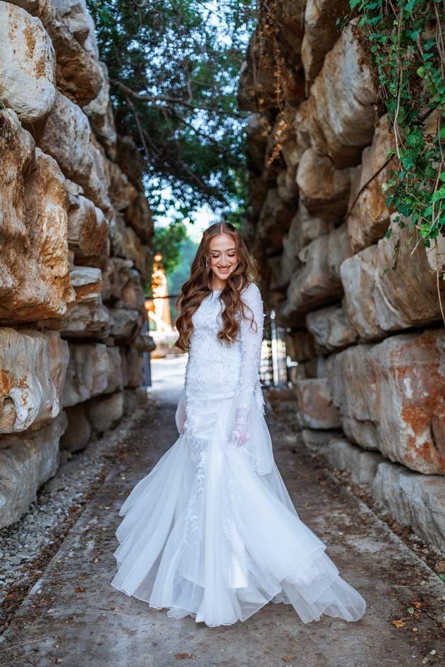 אישה בשמלה לבנה מחייכת, עומדת בין חומות אבן גדולות; שביל מוביל אל רקע עם ירק.