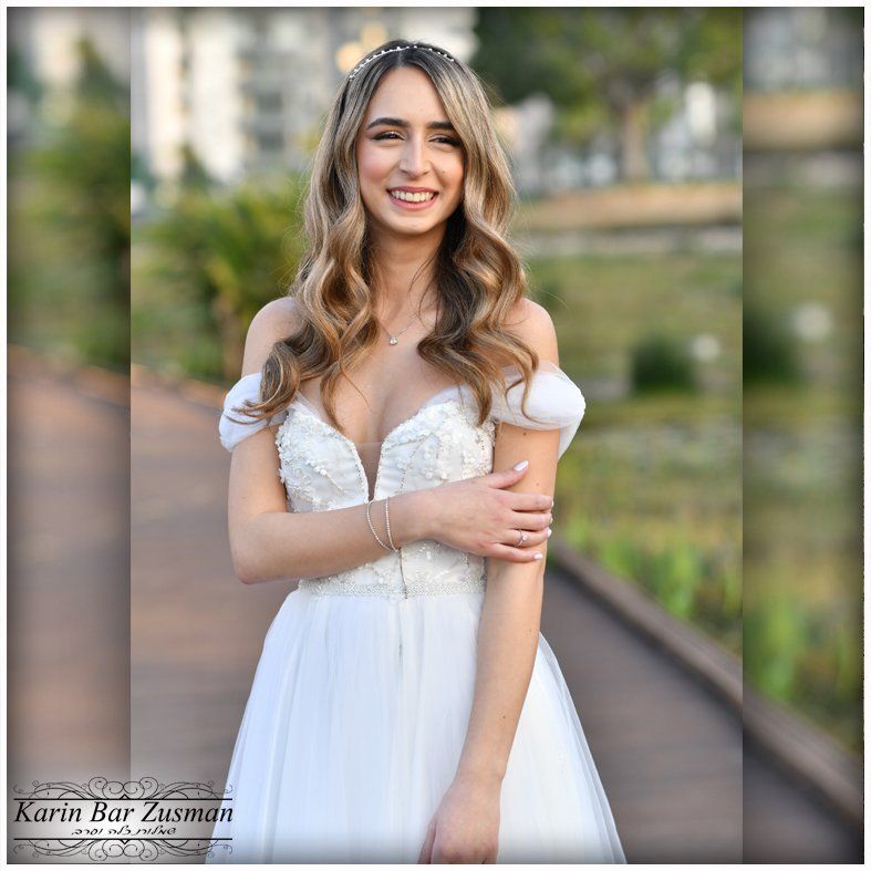 כלה בשמלה לבנה מחייכת בחוץ, שיער גלי, מחוך פרחוני, שרוולים חשופים מהכתפיים, איפור קל.