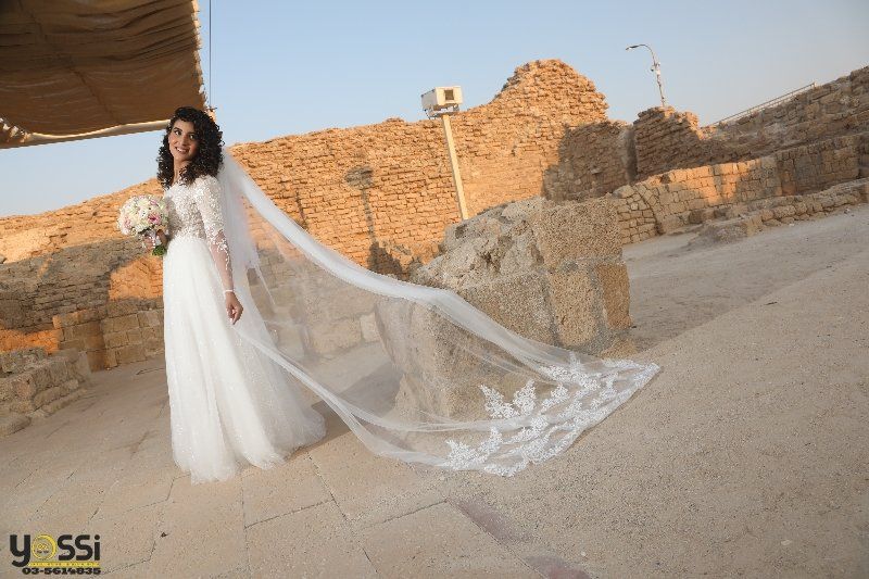 כלה בשמלה לבנה ורעלה מצטלמת בחורבות עתיקות, אוחזת זר, יום שטוף שמש.