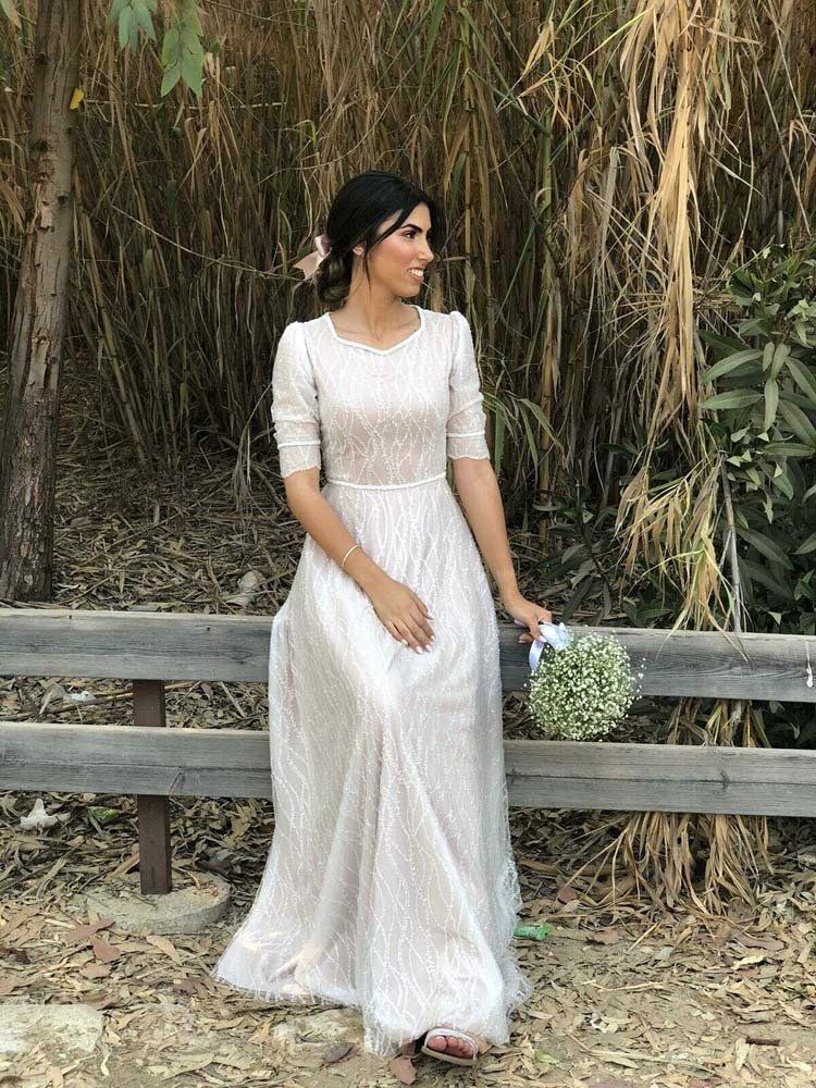 אישה בשמלה לבנה יושבת על ספסל, אוחזת זר פרחים. היא מחייכת על רקע טבעי של חוץ.