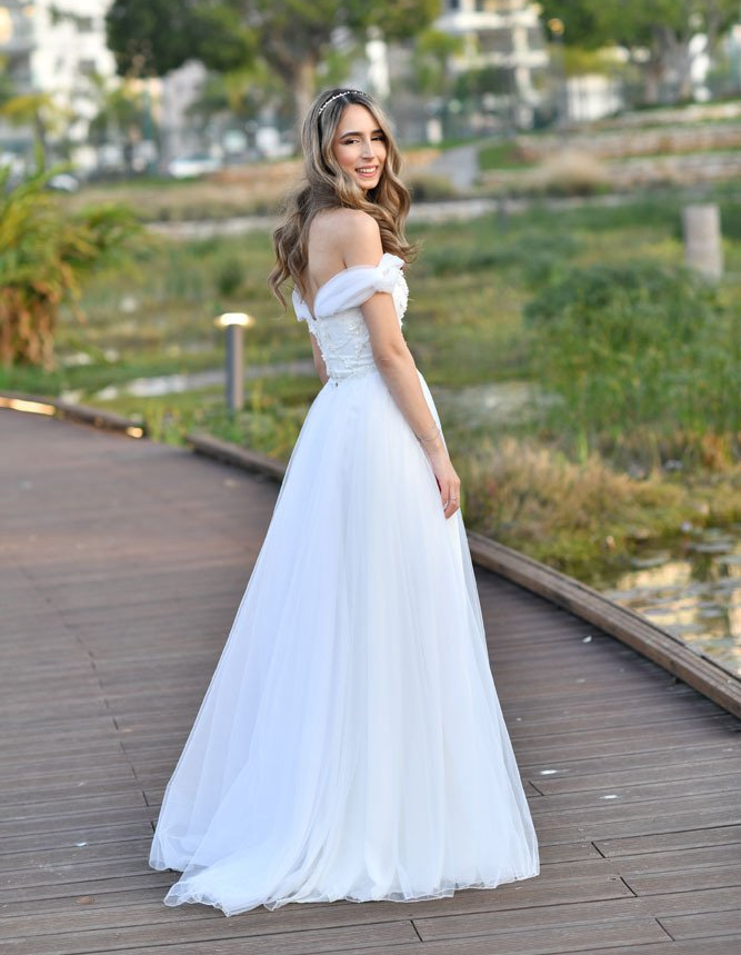 אישה בשמלה לבנה ללא כתפיים מחייכת על טיילת עץ ליד מים.