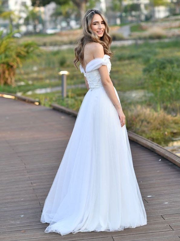 אישה בשמלה לבנה ללא כתפיים מחייכת בחוץ על שביל עץ.
