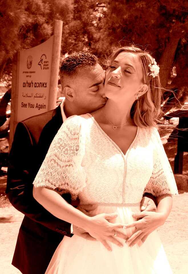 החתן מנשק את צוואר הכלה; היא מחייכת. הם מתחבקים בגווני ספיה בחוץ, ליד שלט.