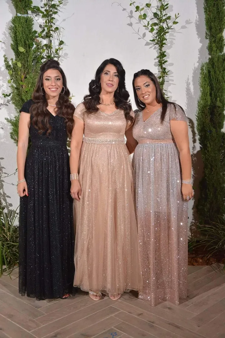 שלוש נשים בשמלות חגיגיות מצטלמות מול צמחייה ירוקה; אחת בשחור, אחת בזהב ואחת בוורוד.