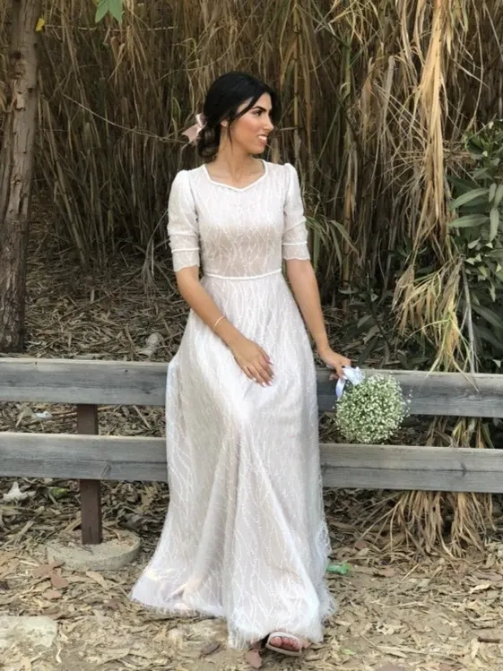 אישה בשמלה לבנה יושבת על ספסל, אוחזת זר פרחים. סביבה חיצונית עם עצים וצמחייה.