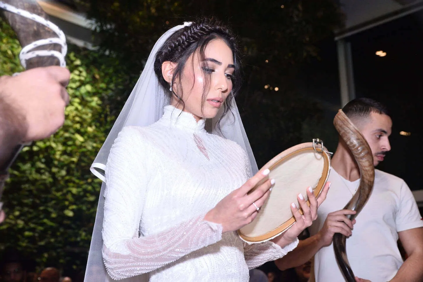 כלה אוחזת בתוף מרים במהלך טקס חתונה. היא לבושה בשמלה לבנה עם רעלה. גבר אחר אוחז בשופר.