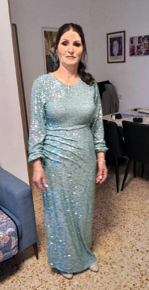אישה בשמלה ארוכה, טורקיז, משובצת פאייטים עומדת בתוך הבית. היא מחייכת חיוך קל.