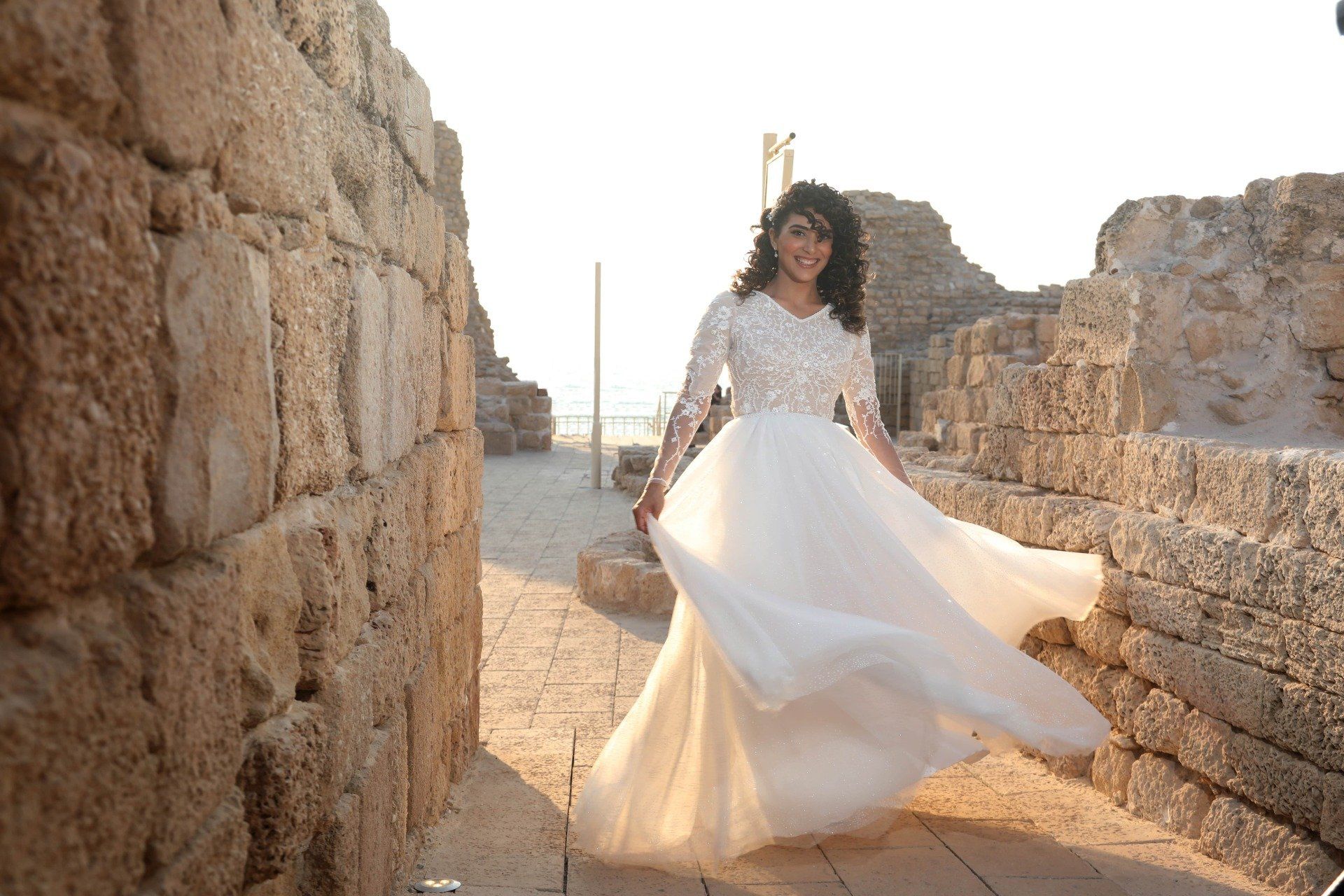 אישה בשמלה לבנה מסתובבת, מחייכת. סביבה של חורבות אבן עתיקות.
