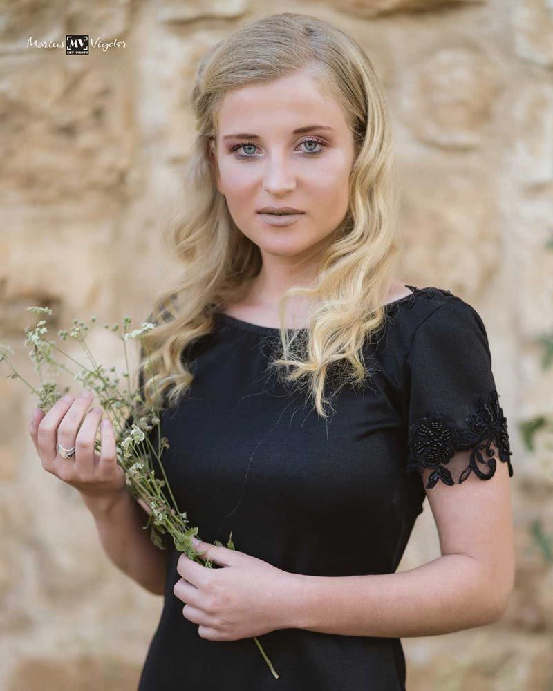 אישה בלונדינית בשמלה שחורה מחזיקה פרחי בר, עומדת ליד קיר אבן ומביטה בצופה.
