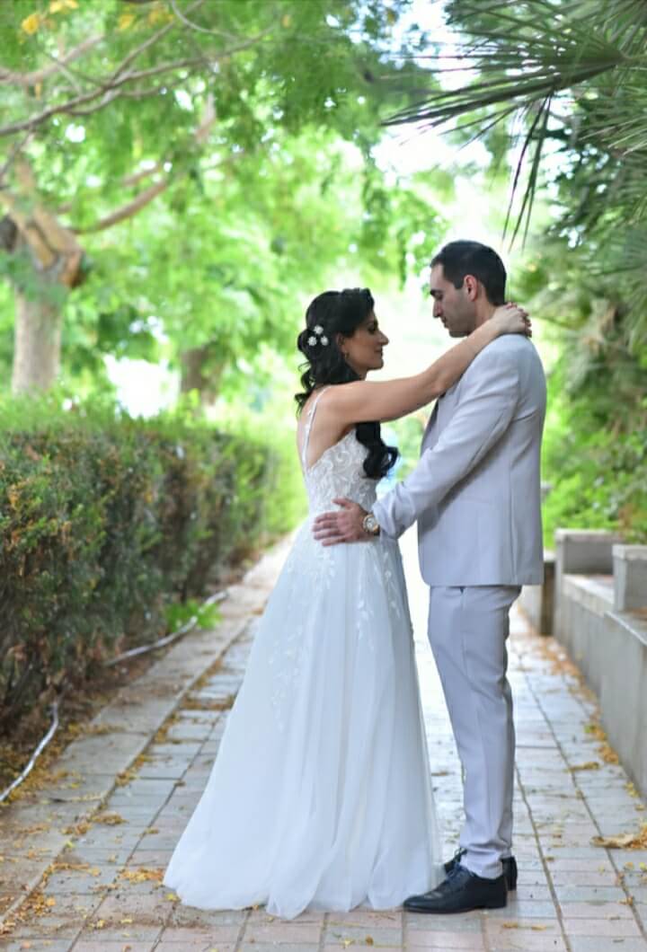 חתן וכלה מתחבקים על שביל לבנים מוקף בירק. היא לובשת שמלה לבנה, הוא לובש חליפה אפורה.