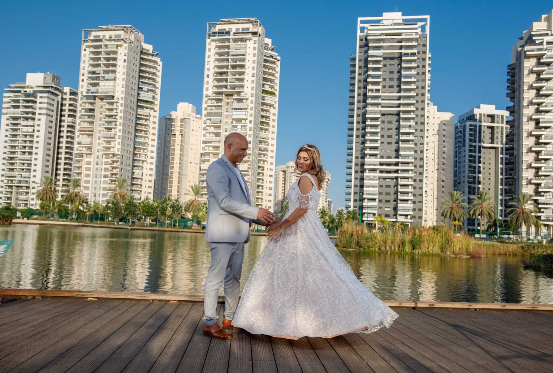 חתן וכלה מחזיקים ידיים על דק עץ ליד אגם, כשברקע גורדי שחקים של העיר.