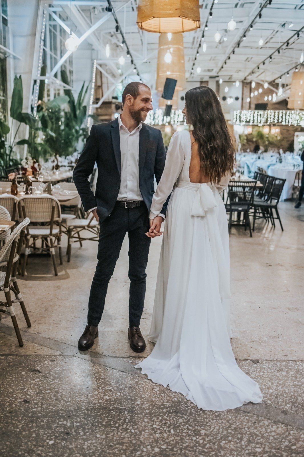 זוג חתן אוחז ידיים ומחייך. גבר בחליפה, אישה בשמלת כלה לבנה; סביבה פנימית.