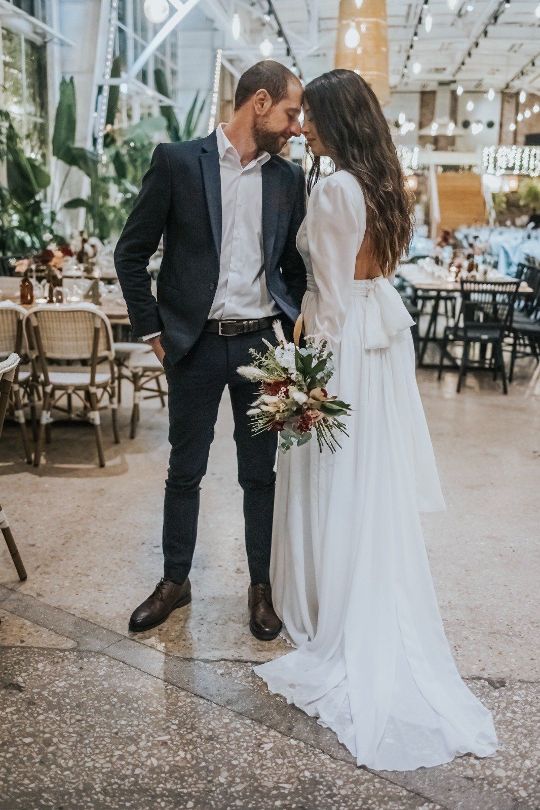 חתן וכלה נוגעים במצחם, אוחזים בפרחים. הם עומדים בתוך הבית.
