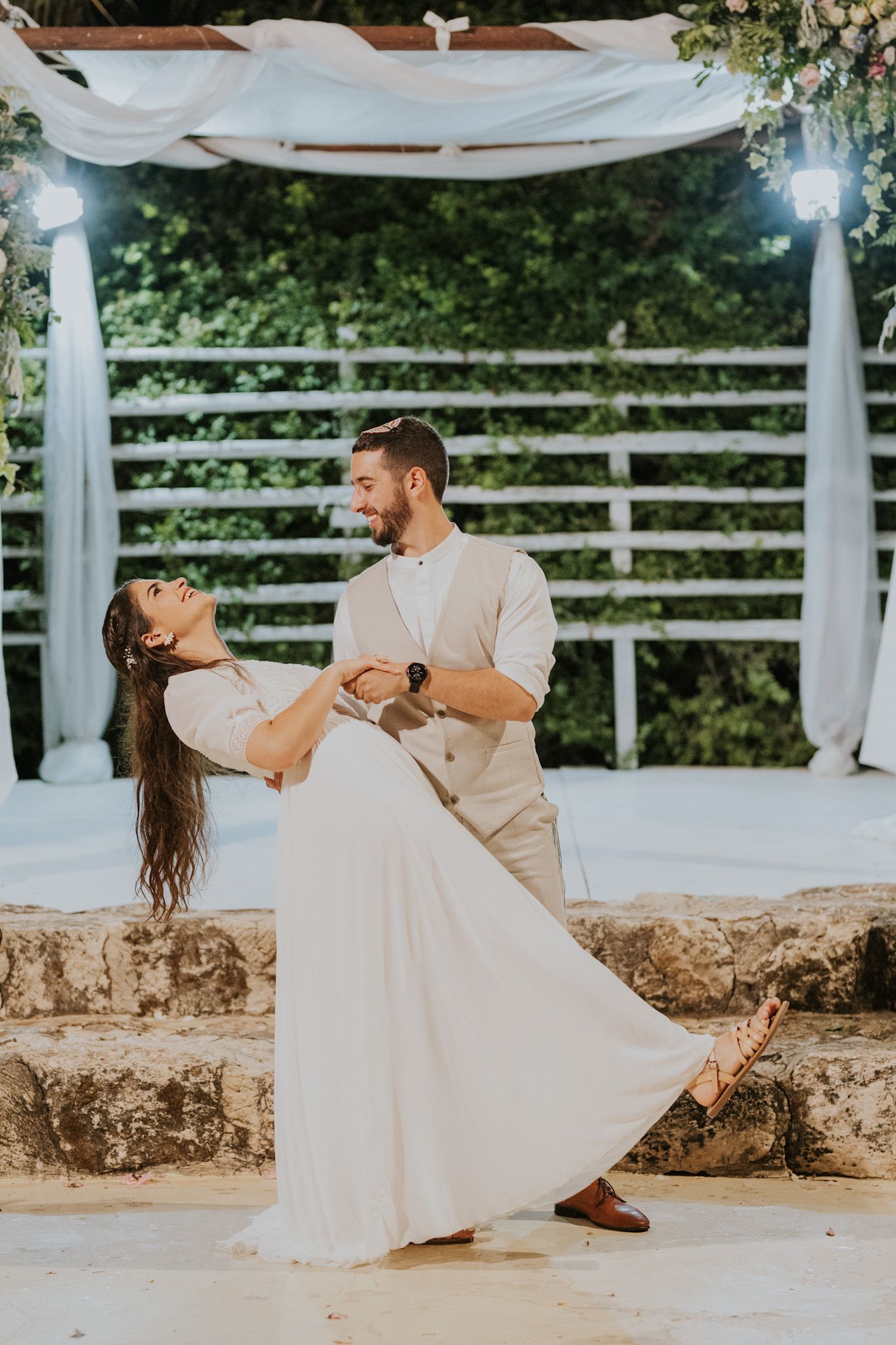 חתן וכלה רוקדים על פטיו אבן מתחת לקשת, הוא בגופיה, היא בשמלה לבנה, מחייכים.