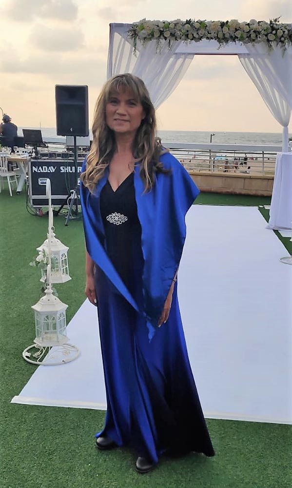 אישה בשמלה כחולה וצעיף עומדת באירוע חיצוני; קשת לבנה ואוקיינוס נראים לעין.