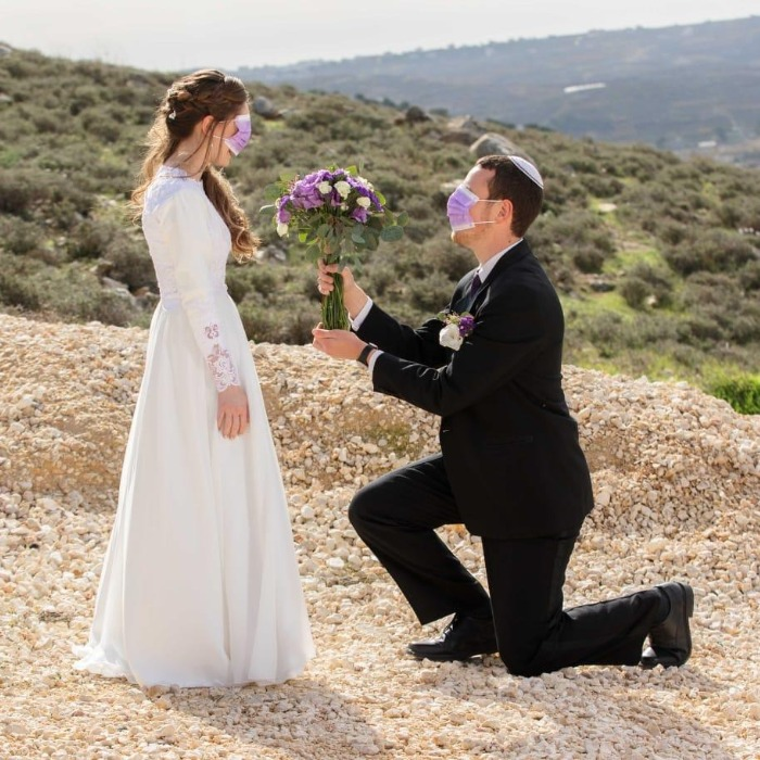 כלה בשמלה לבנה וחתן בחליפה כורעים ברך, שניהם עונדים מסכות, בחוץ, החתן מציע פרחים.