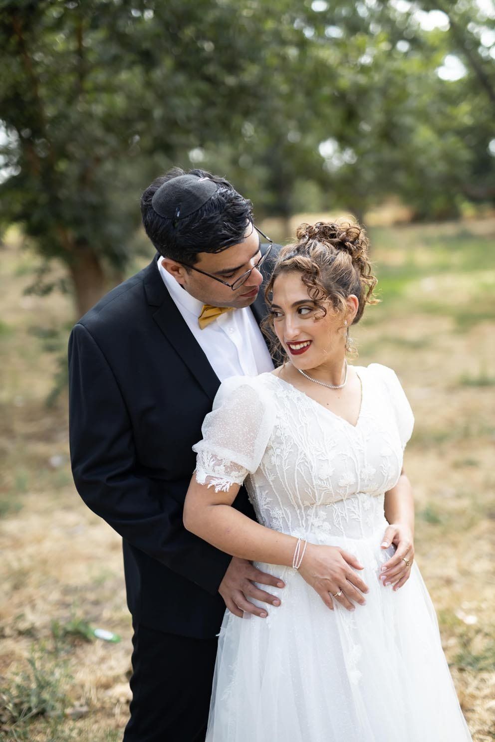 חתן בחליפה שחורה מנשק את לחיה של הכלה. כלה בשמלה לבנה מחייכת בחוץ.