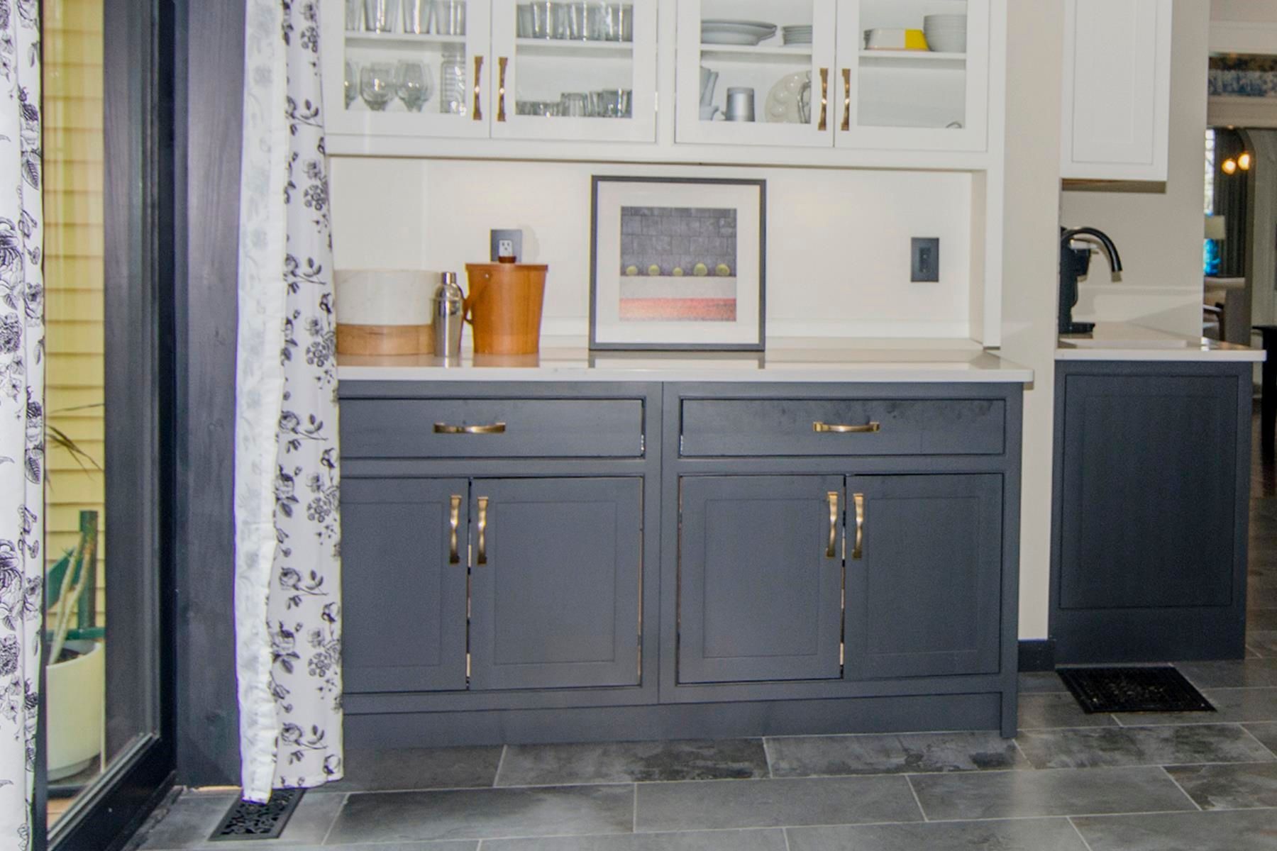 Dark blue cabinets with brass hardware, white countertop, white upper cabinets, artwork, and curtains.
