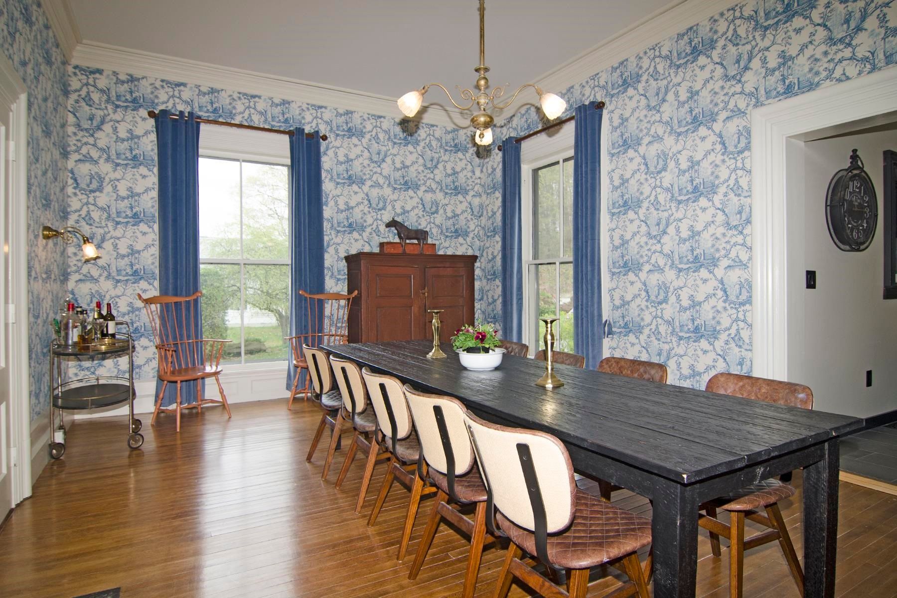 Dining room with blue patterned wallpaper, long black table, brown chairs, and wooden floor.