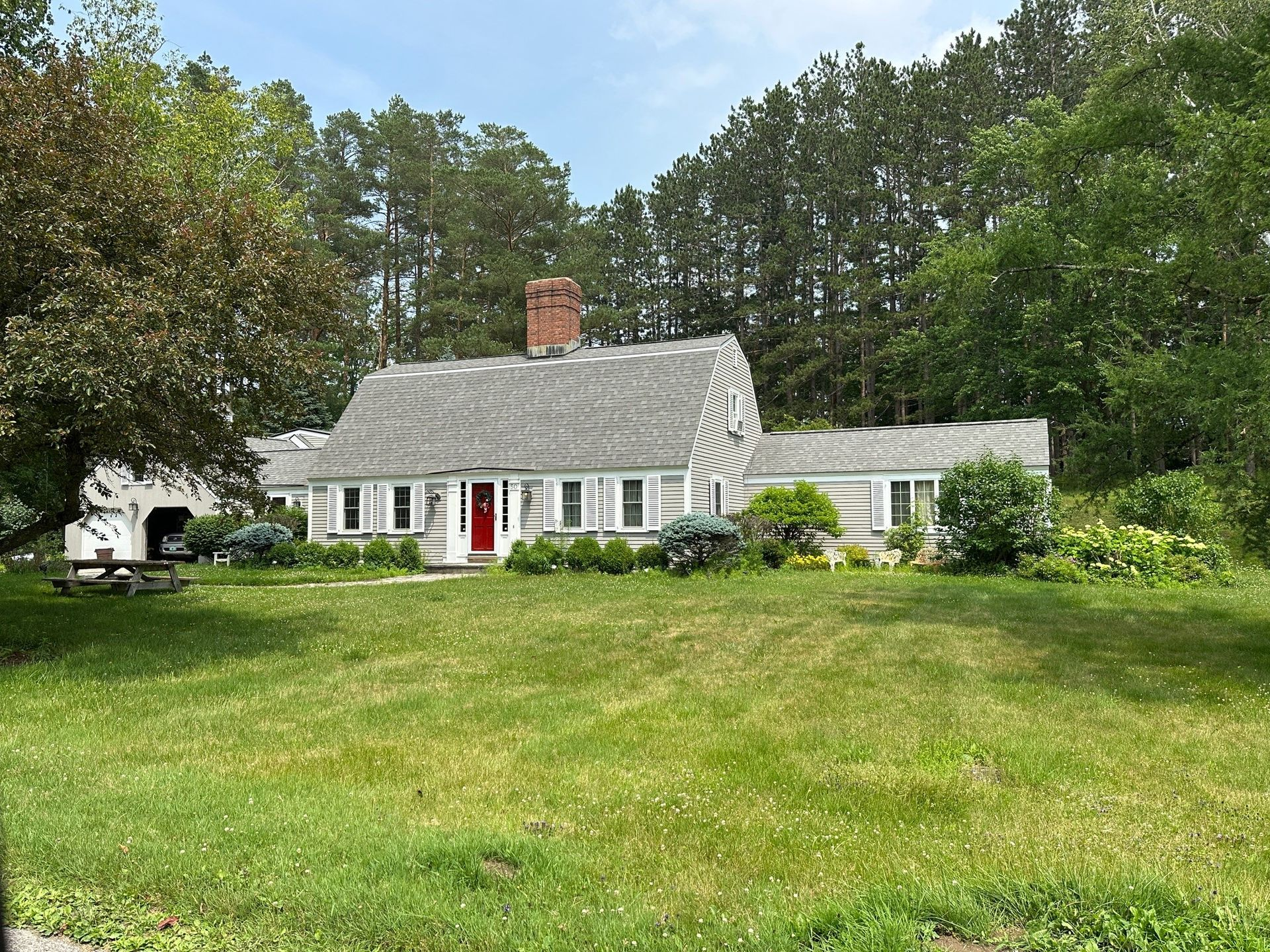 Cottage with a gray shingle roof, red door, and brick chimney set among trees and a green lawn.