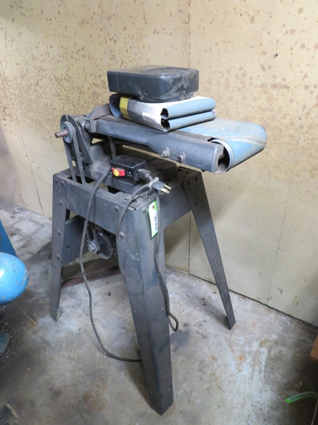 A black belt sander on a metal stand in a workshop. The sander is gray with a black electrical cord.