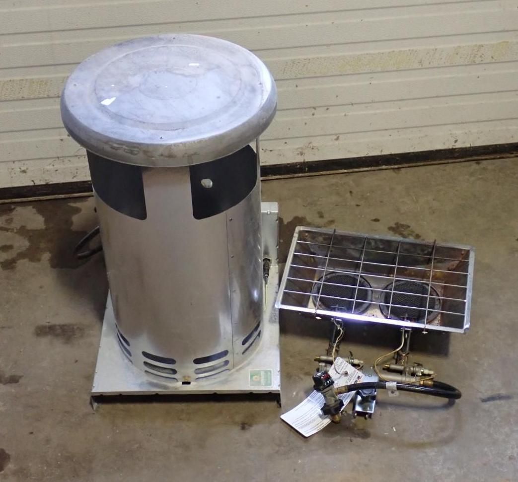 A silver cylindrical space heater stands next to a detached, two-burner metal camping stove on a concrete floor.