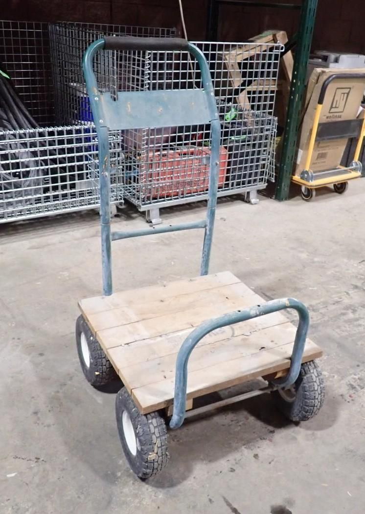 A small, four-wheeled utility cart with a wooden platform and a tall blue metal handle, sitting on a warehouse floor.