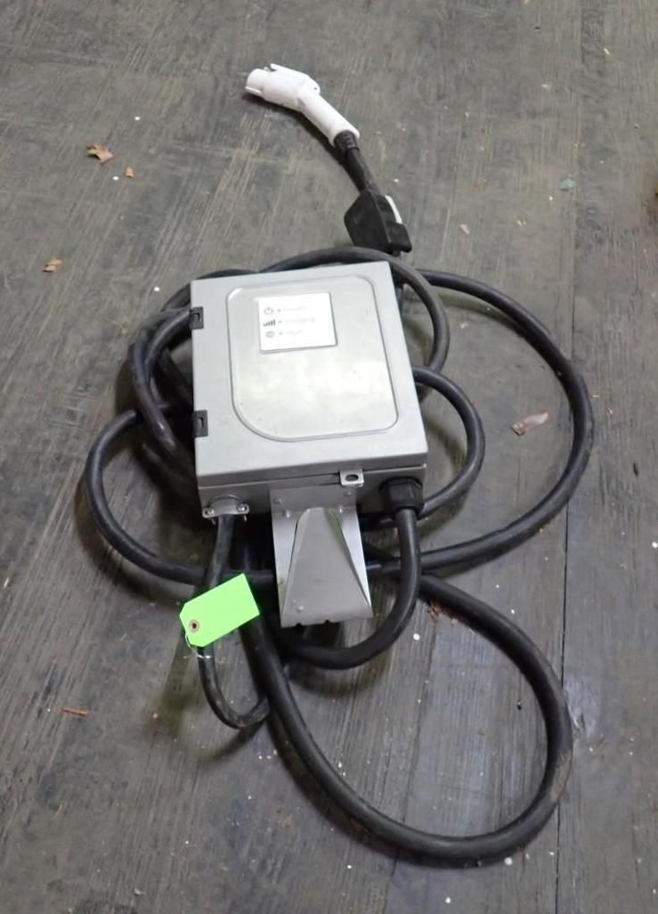 A gray EV charging station unit with a black charging cable and white connector plug resting on a dark floor.