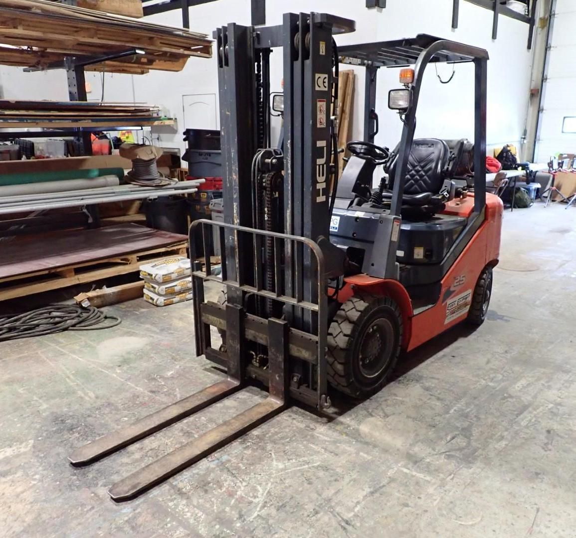 A red Heli forklift parked in a warehouse, with its metal forks extended on a concrete floor near storage shelves.