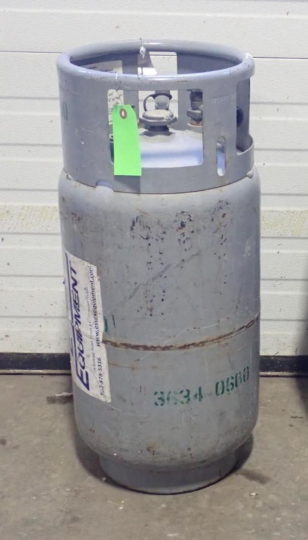A grey industrial gas cylinder with a protective collar stands on a concrete floor against a white paneled wall.